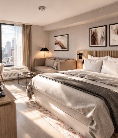 A modern hotel room is furnished with a bed, desk, coffee maker, and sofa, bathed in natural light, offering a city skyline view from a large window.