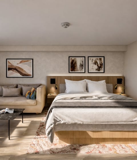A neatly made bed and beige sofa furnish a modern hotel room. The bed has white pillows and a textured rug lies underneath. A wall display shows "25°".