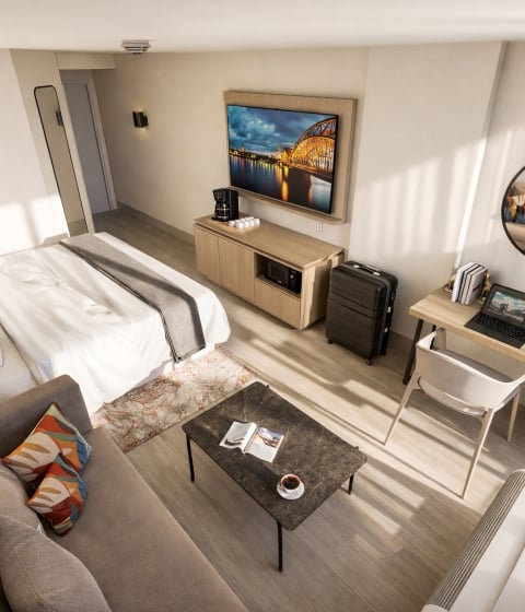 A modern hotel room features a bed, sofa, workstation with laptop, and TV showing a night bridge. Sunlight fills light wood and white walls; a thermostat reads "20".