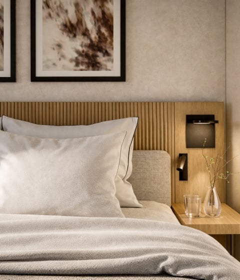 A neatly made bed with light pillows and blankets features a wooden headboard, abstract art, and a nightstand with a glass and vase, within a sunlit, modern bedroom.
