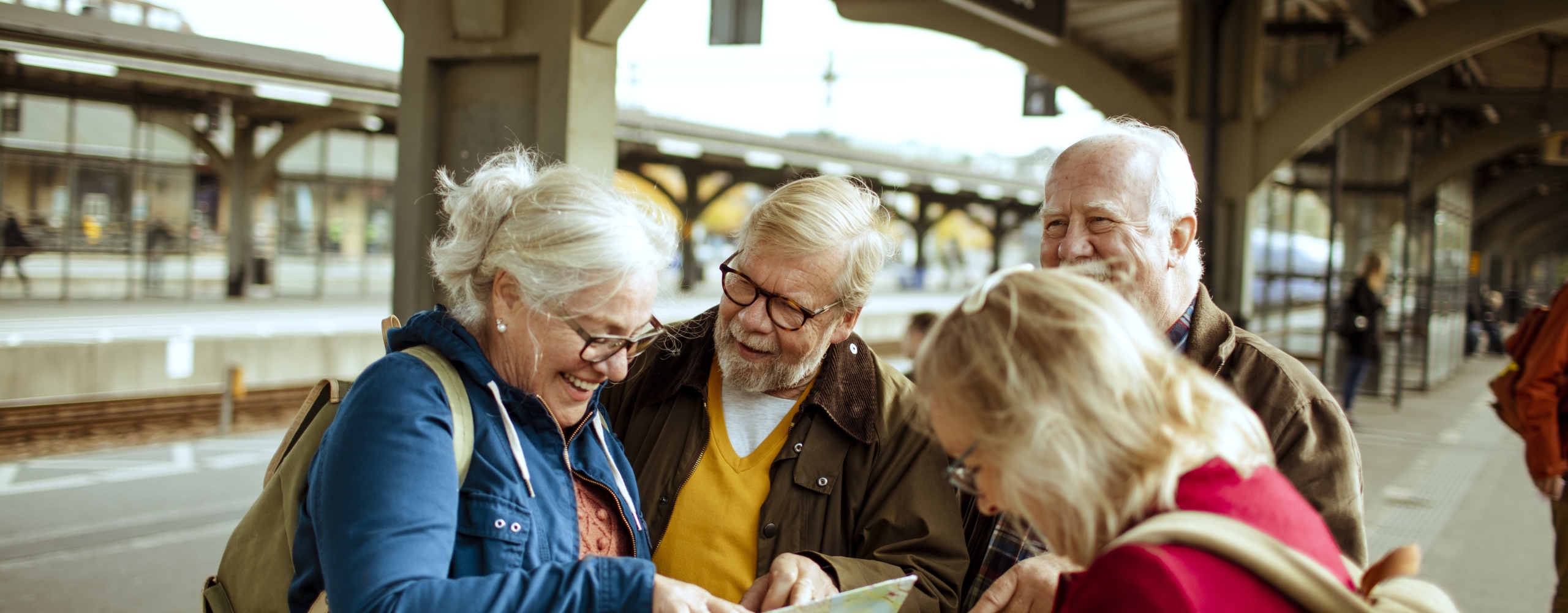 A group of four senior travelers smiling and looking at a paper map together on a train platform.