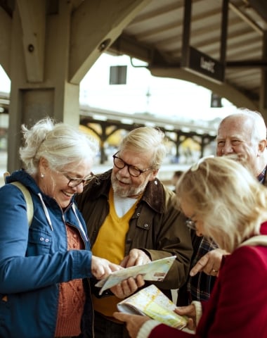A group of four senior travelers smiling and looking at a paper map together on a train platform.