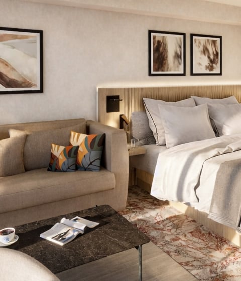A modern hotel room features a king bed, bedside tables, and a beige sofa with colorful cushions. Sunlight illuminates the room's warm, minimalist decor.