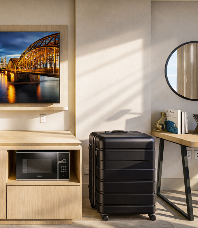 A black rolling suitcase stands upright next to a light wood cabinet housing a "BOSCH" microwave, below a TV displaying an illuminated bridge scene in a sunlit room.