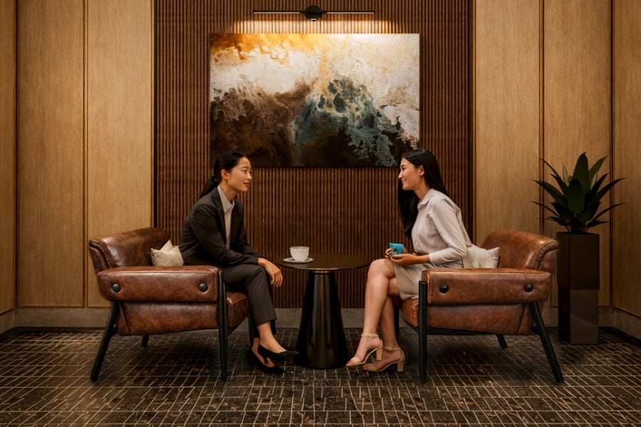 Two women converse while seated in brown lounge chairs facing each other, separated by a small table with a cup, in a modern wood-paneled room with an abstract painting.