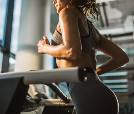 A sweaty woman runs on a treadmill, viewed from the side. Her arms are bent, and she wears a sports bra and leggings in a brightly lit gym.