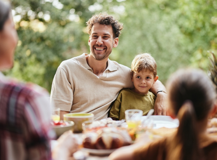 A smiling man with his arm around a young boy sits at an outdoor dining table filled with food. Other blurred family members are also present.