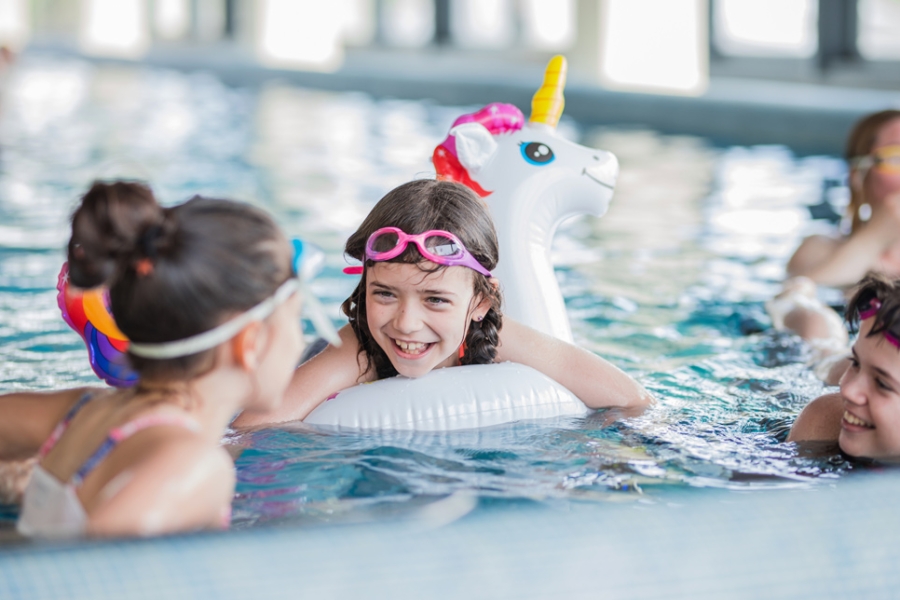 A young girl with pink goggles smiles broadly while resting on a white inflatable in a swimming pool with other children and an inflatable unicorn toy.