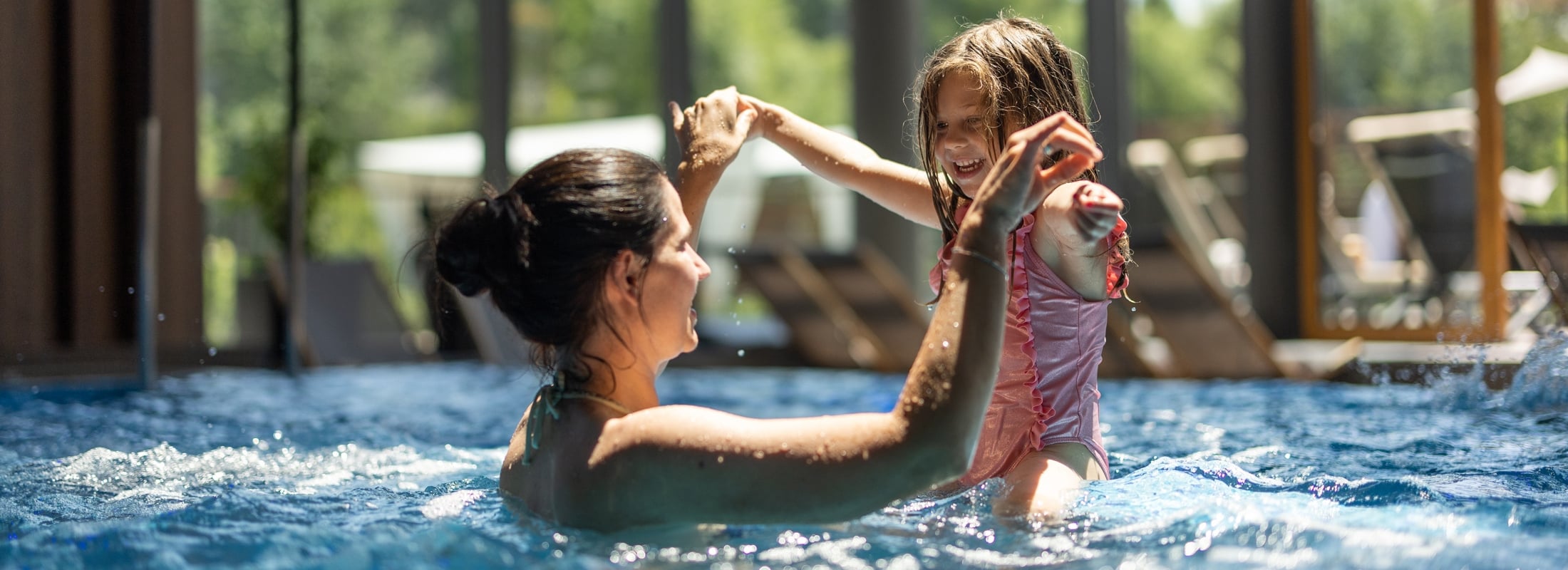 A woman and a young girl play joyfully in a sunny pool, holding hands and smiling. Large windows reveal lush green hills and sun loungers in the background.