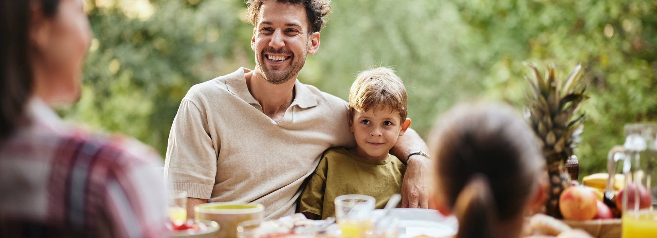 A smiling man has his arm around a boy, seated at an outdoor table with food and drinks. Other blurry people are present amidst green foliage.