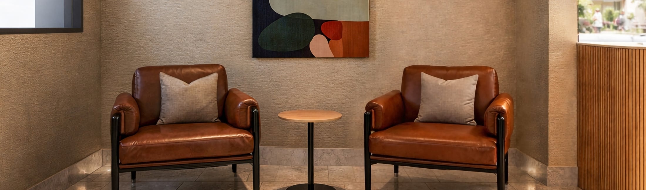 Brown leather armchairs with pillows face forward, separated by a round wooden side table. They're on a tiled floor in a modern lounge with textured beige walls and an abstract painting.