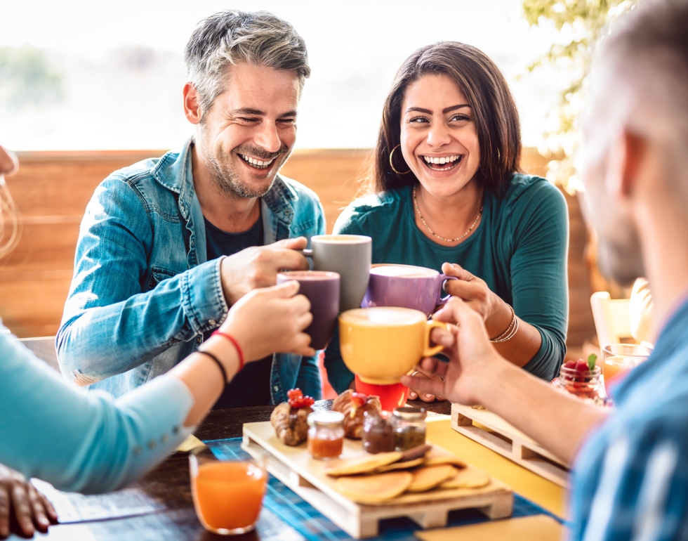 Four diverse friends laugh while clinking colorful coffee mugs together at a table laden with breakfast pastries, pancakes, and juice on an outdoor patio.