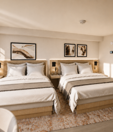 Two queen-sized beds are neatly made with neutral bedding and throw blankets in a modern hotel room featuring warm lighting, framed art, and a patterned rug.