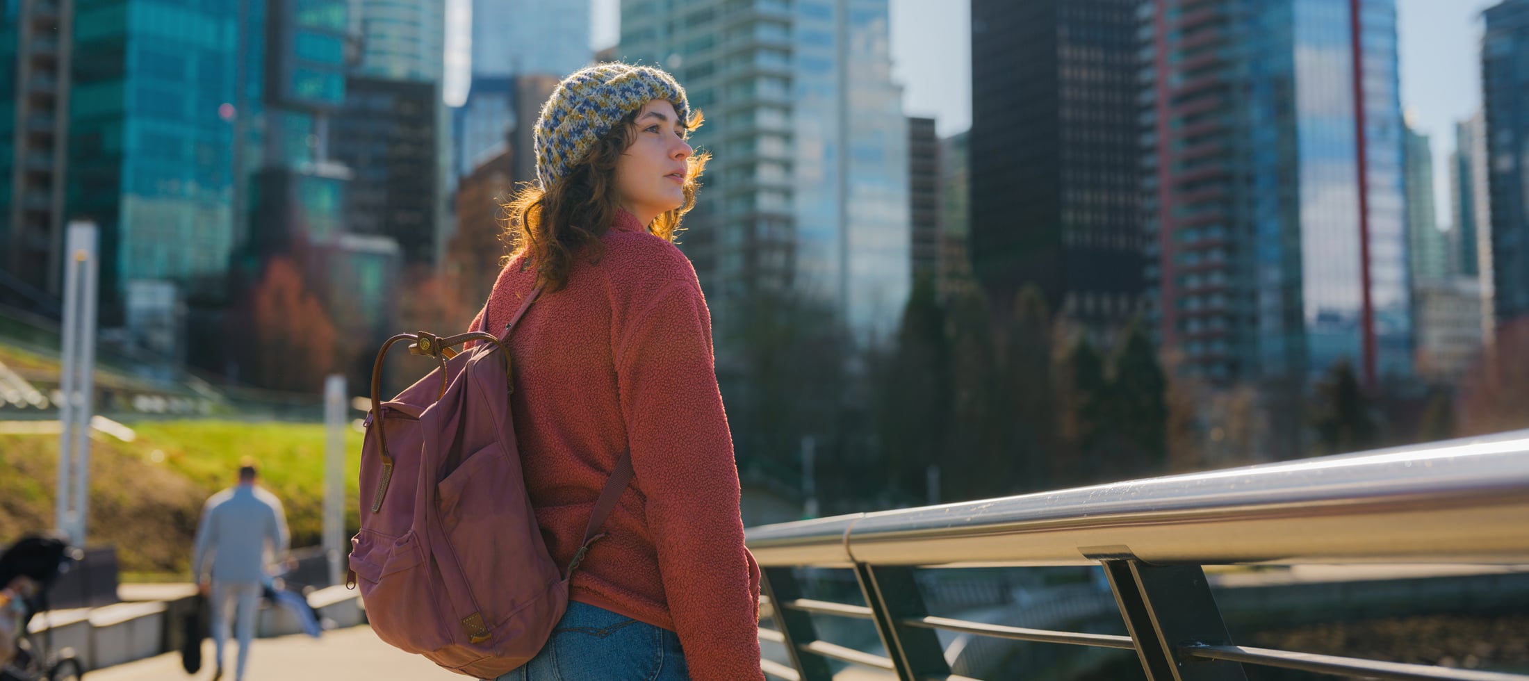 A young woman in a beanie and red fleece, wearing a purple backpack, looks right, backlit by sun. She stands by a railing with city skyscrapers blurred behind her.