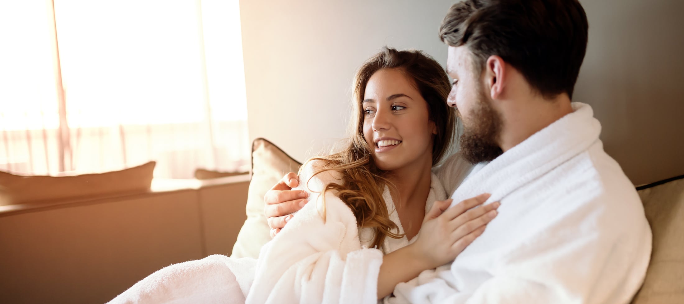 A smiling woman in a white bathrobe embraces a man wearing a matching robe, looking at him warmly indoors with bright window light in the background.