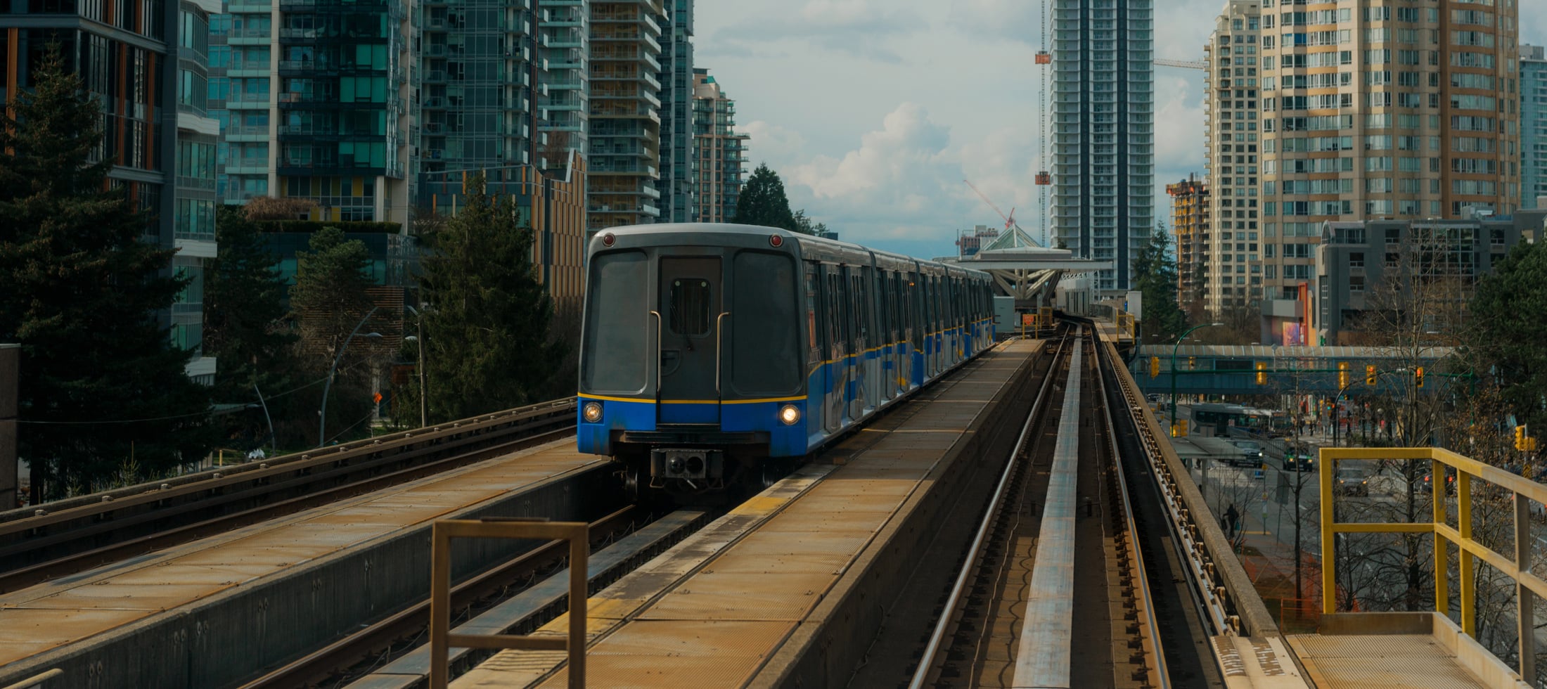A grey and blue train travels on elevated tracks through a modern city skyline with tall buildings and trees under a cloudy sky.