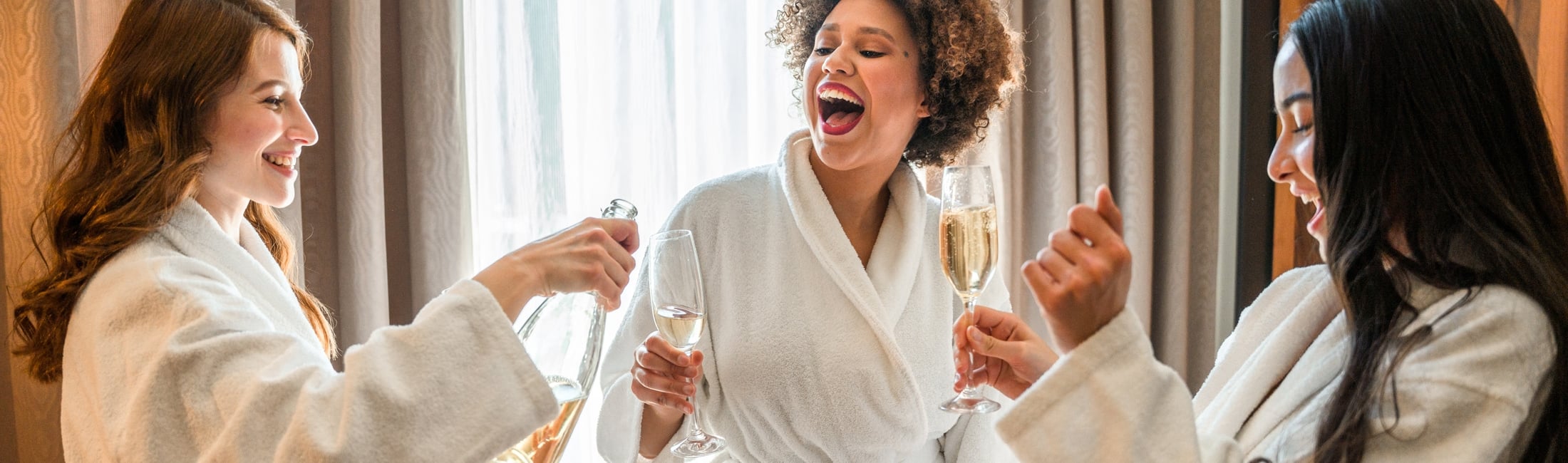 Three women in white bathrobes are laughing and smiling, one pouring champagne into a glass held by another, indoors near a window with curtains.