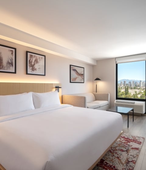 Hotel room with a king bed, framed art above, and soft lighting; large window offers a city skyline view.