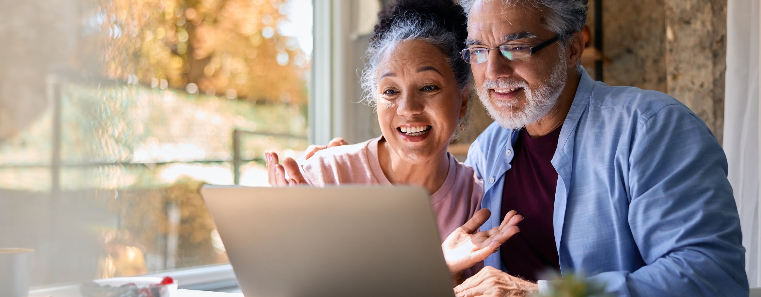 A happy senior couple looks intently at a laptop, with the woman gesturing, while sitting at a table by a sunlit window revealing autumn trees.