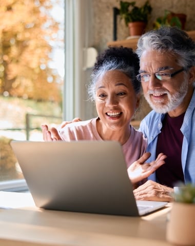 A happy senior couple looks intently at a laptop, with the woman gesturing, while sitting at a table by a sunlit window revealing autumn trees.