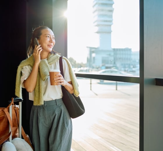 A smiling woman talks on her phone, holding a coffee cup. She stands by a bright window overlooking an airport terminal, with luggage next to her.