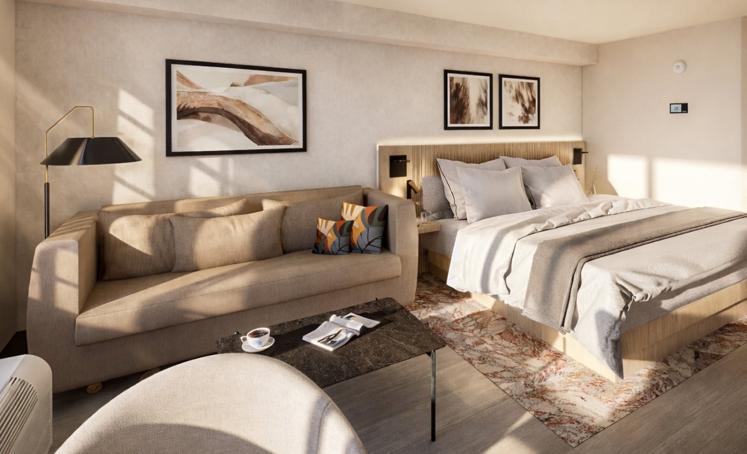 A modern studio room features a large bed and a beige sofa. Sunlight streams onto a coffee table with a magazine and cup, illuminating the light-toned space.