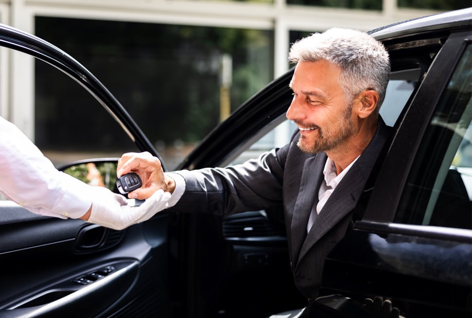 A smiling man in a suit, seated in a black car, hands car keys to a white-gloved person's hand, likely for valet service.