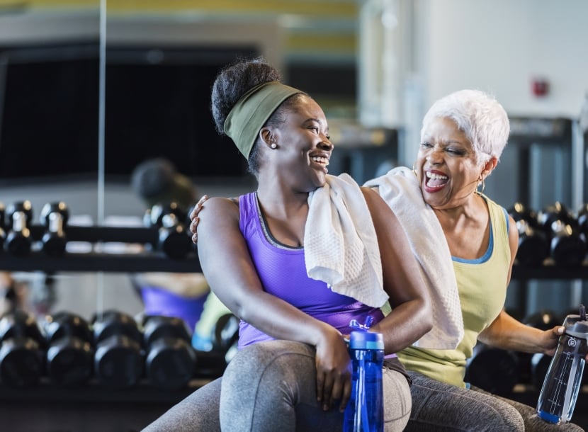 A senior African-American woman in her 60s exercising at the gym with her adult daughter, a young woman in her 20s. They are taking a break from lifting dumbbells, conversing and drinking from water bottles.