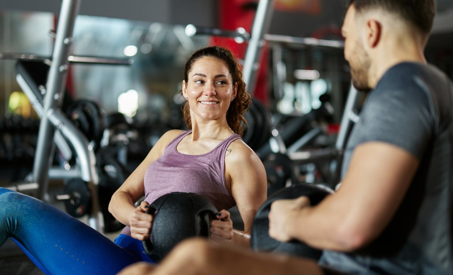 A smiling woman holds a medicine ball, looking at a man as they exercise together on a bench in a gym.