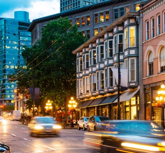 A historic bay-windowed building stands illuminated amidst blurred traffic and glowing streetlights on a city street at dusk, flanked by urban architecture.
