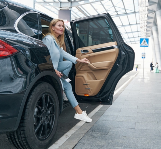 Blonde woman in a blue suit steps out of a black car, holding tickets, looking right. She's at an airport drop-off, near a blue pedestrian crossing sign.