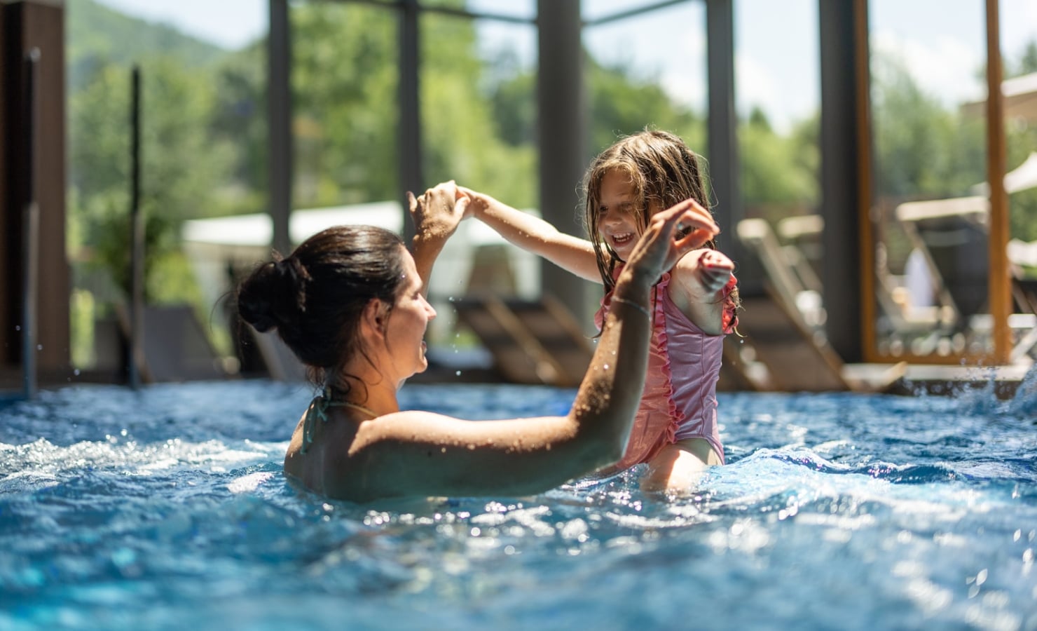 A woman and a young girl hold hands, laughing and splashing in a swimming pool, with large windows overlooking green trees and mountains in the background.