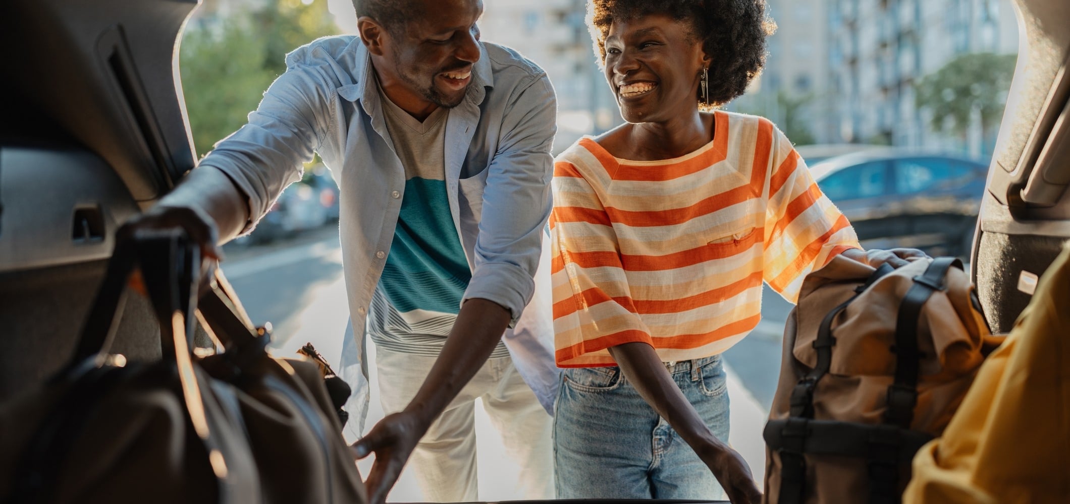 A smiling Black couple loads travel bags into a car trunk, outdoors on a sunny day with city buildings in the background.