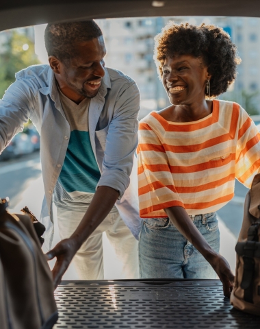 A smiling Black couple loads travel bags into a car trunk, outdoors on a sunny day with city buildings in the background.