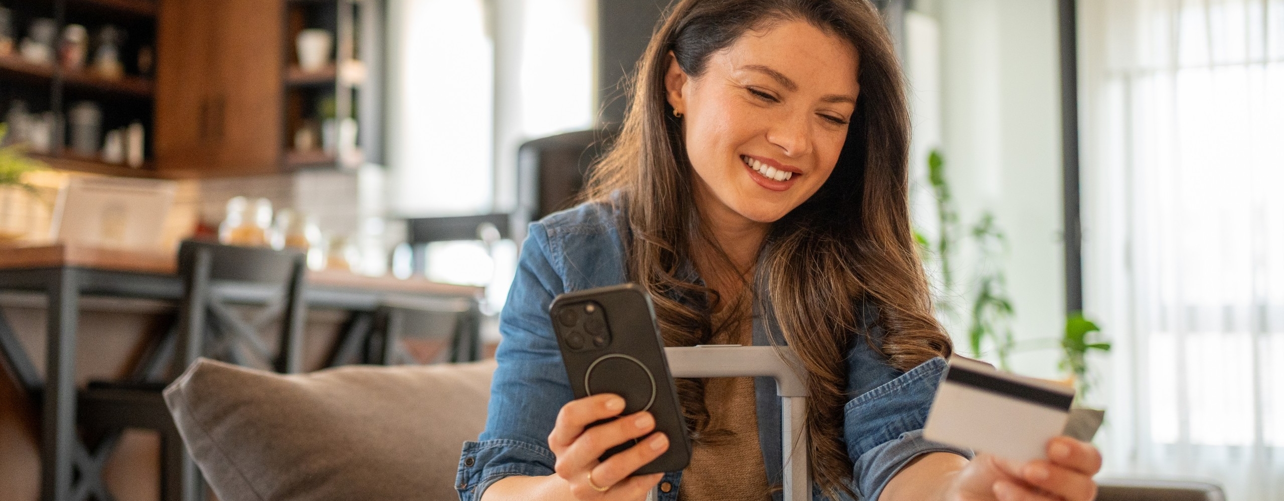 Young woman sitting on the sofa at home, holding her trolley bag and booking a trip online using her smartphone and credit card