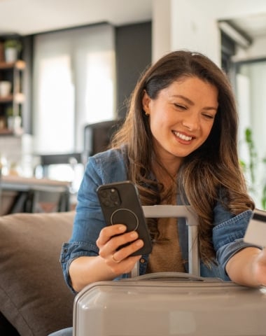 Young woman sitting on the sofa at home, holding her trolley bag and booking a trip online using her smartphone and credit card