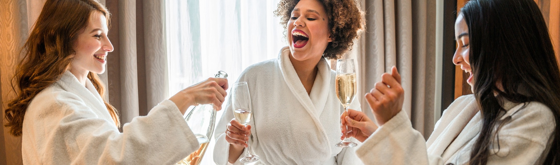 Three women in white bathrobes are laughing and smiling, one pouring champagne into a glass held by another, indoors near a window with curtains.