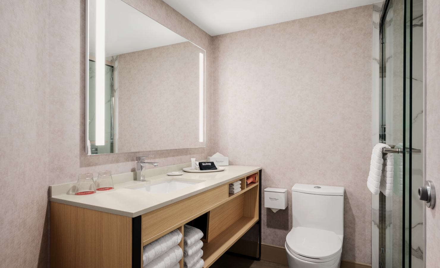 Modern bathroom with a wooden vanity and white countertop, a large backlit mirror, and a glass-enclosed shower to the right; towels stacked on open shelves beneath the sink area.