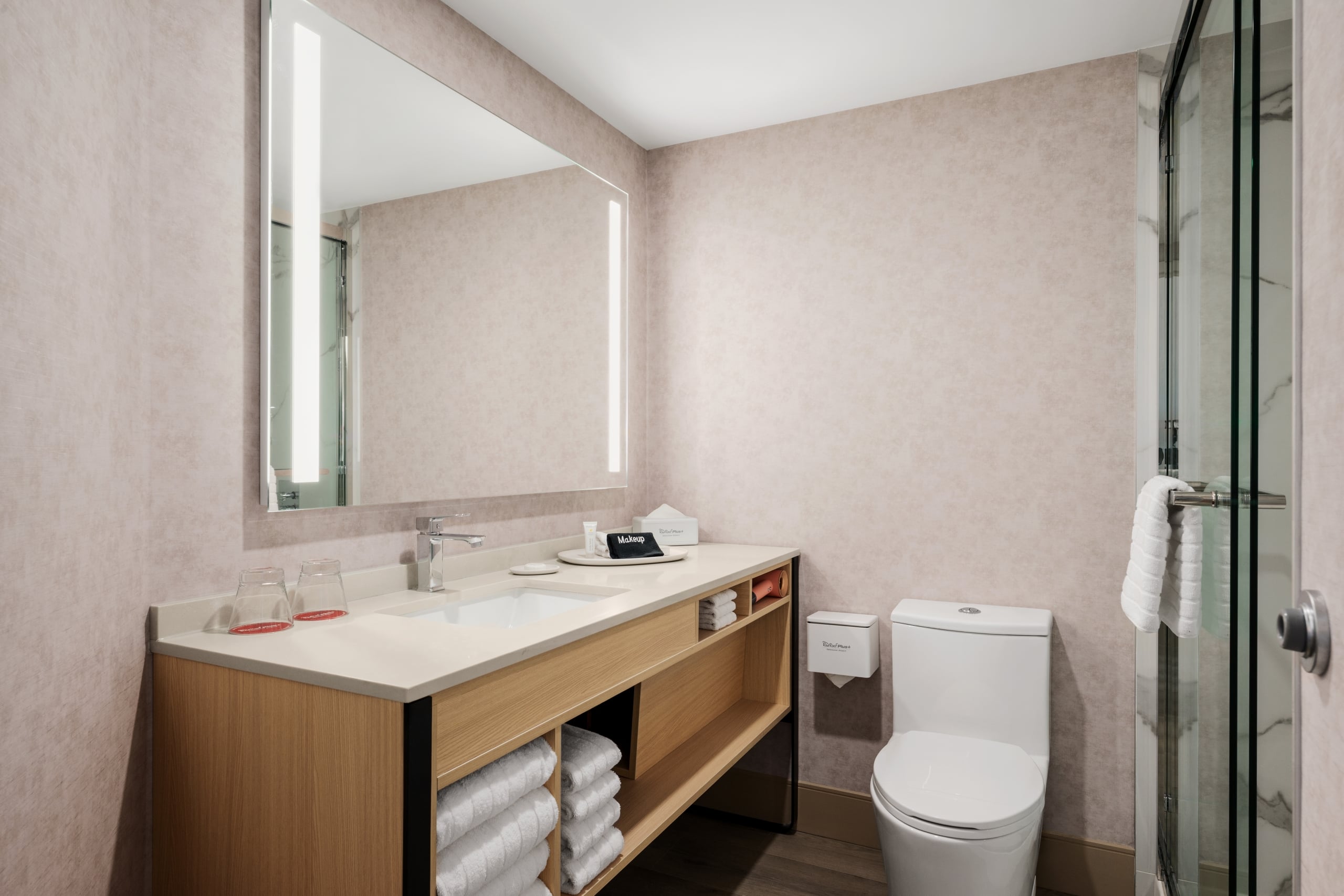 Modern bathroom with a wooden vanity and white countertop, a large backlit mirror, and a glass-enclosed shower to the right; towels stacked on open shelves beneath the sink area.