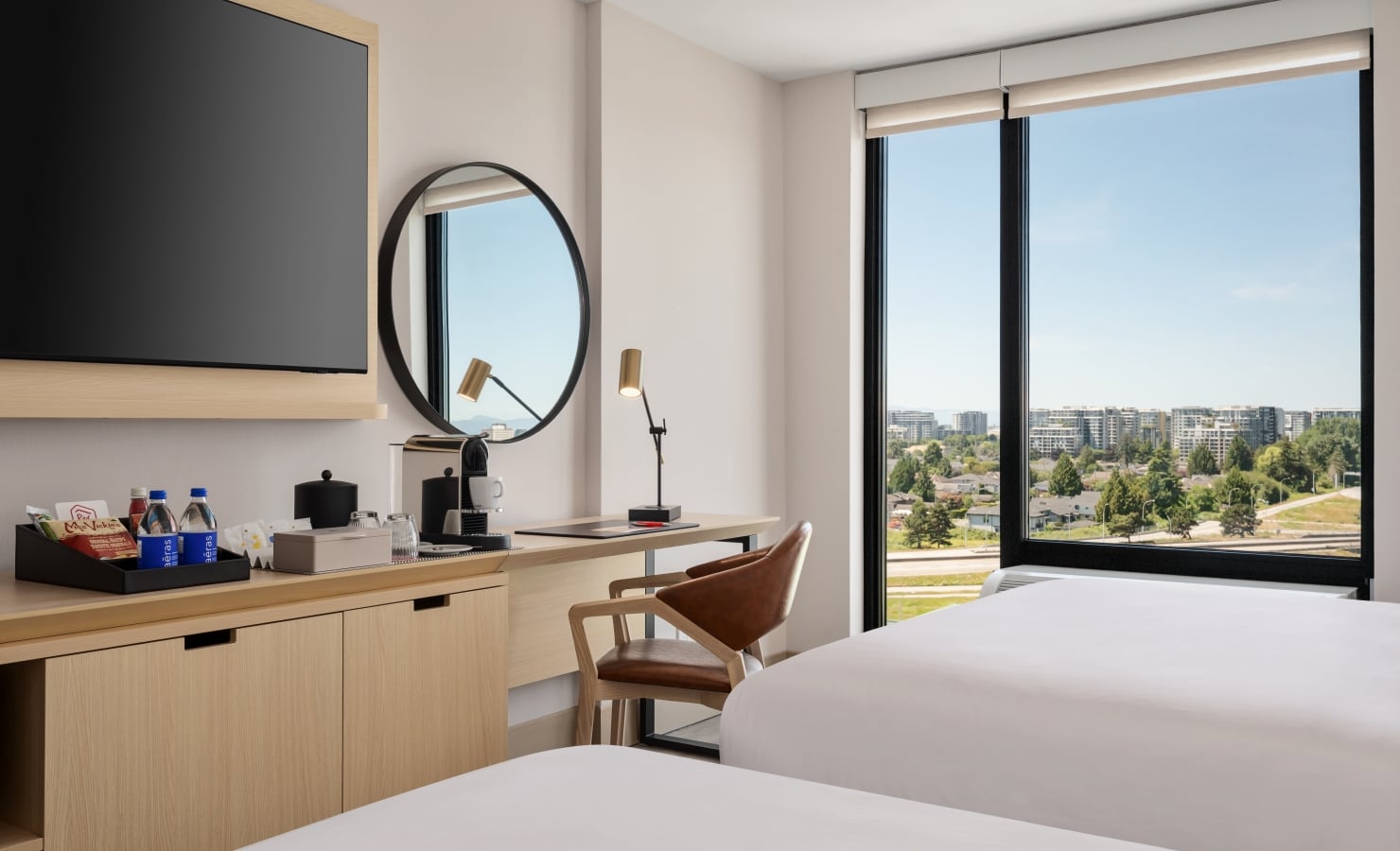 Hotel room workspace: wooden desk with a coffee station, snacks, and a round mirror, plus a large window with a city view.