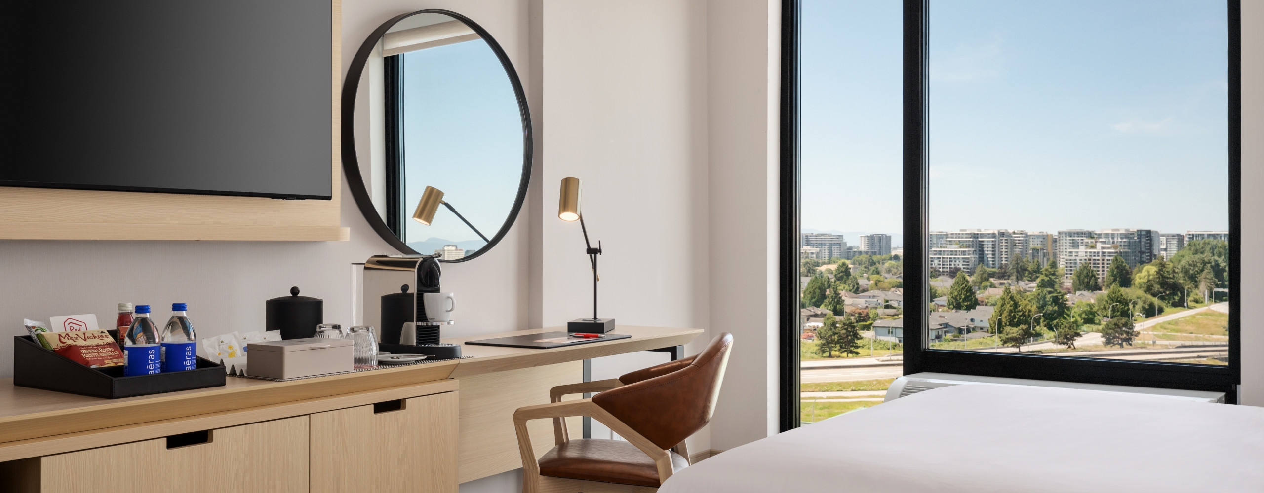 Hotel room workspace: wooden desk with a coffee station, snacks, and a round mirror, plus a large window with a city view.