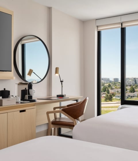 Hotel room workspace: wooden desk with a coffee station, snacks, and a round mirror, plus a large window with a city view.