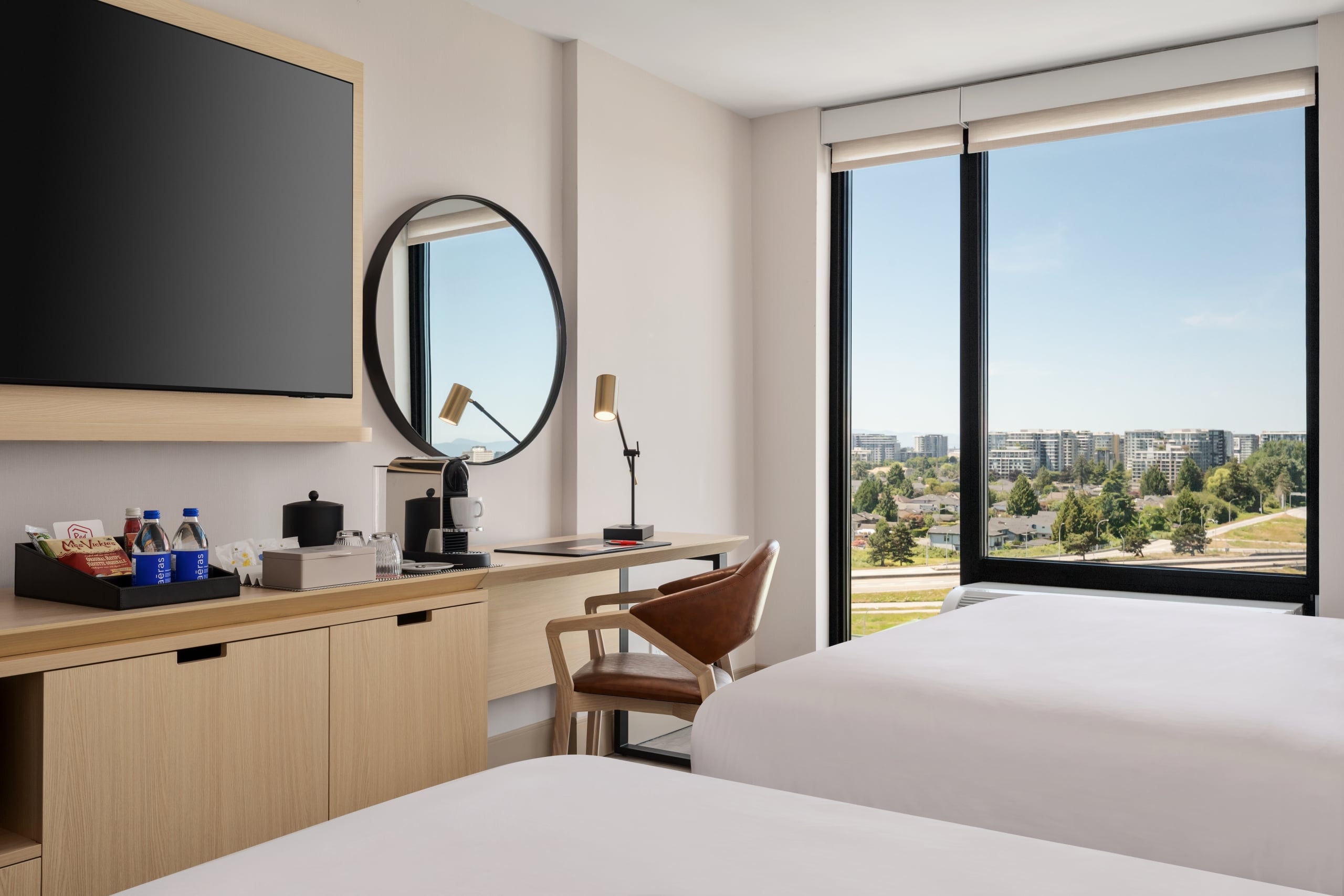 Hotel room workspace: wooden desk with a coffee station, snacks, and a round mirror, plus a large window with a city view.
