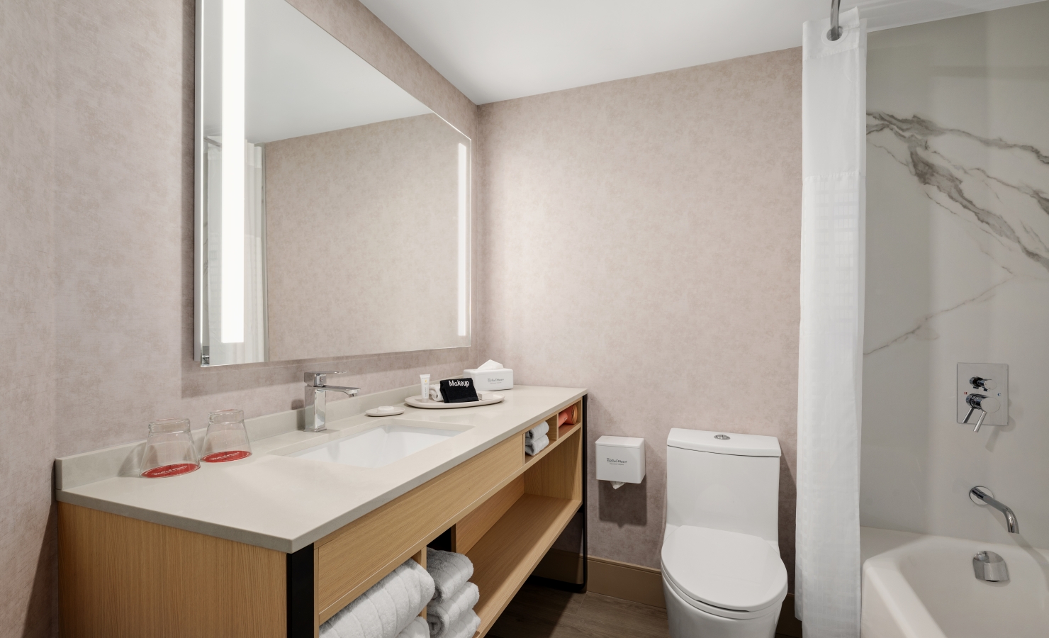 Modern bathroom with a double-sink vanity, large mirror, and open shelving holding white towels; beige textured walls and a white bathtub/shower to the right with a marble-tiled wall.