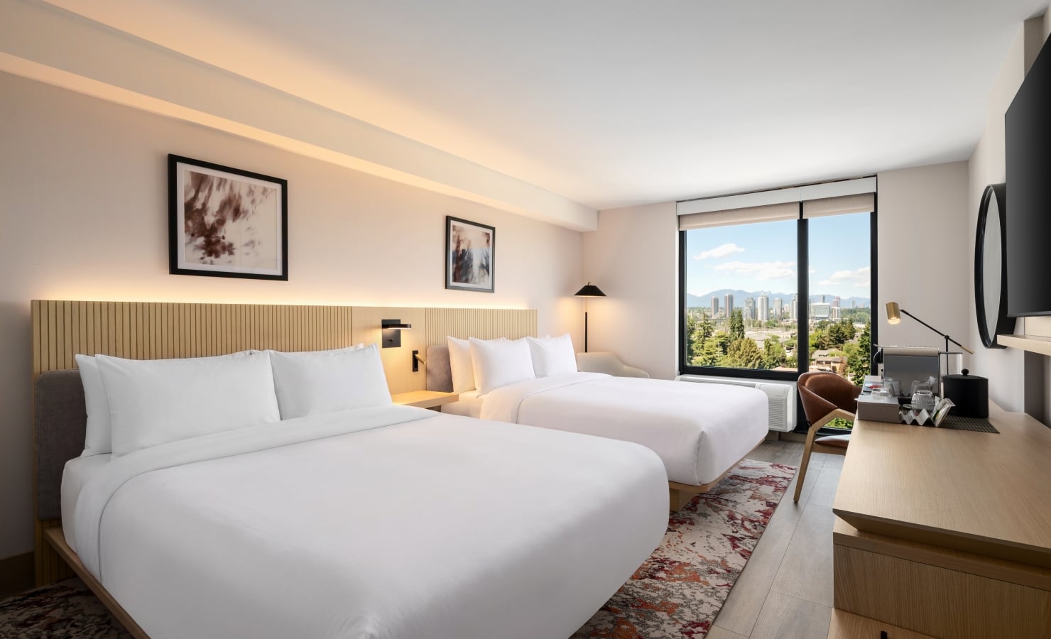 Hotel room with two white-bedding beds, wooden headboard, and a large window overlooking a distant cityscape and hills.