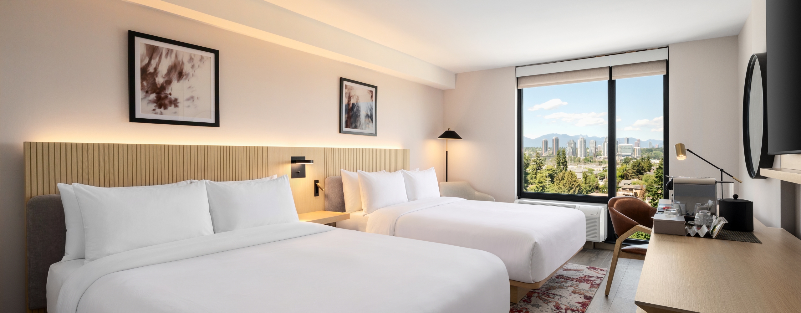 Hotel room with two white-bedding beds, wooden headboard, and a large window overlooking a distant cityscape and hills.