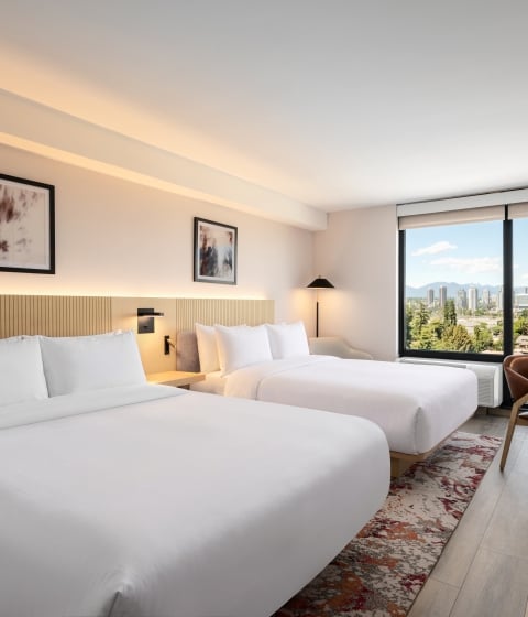 Hotel room with two white-bedding beds, wooden headboard, and a large window overlooking a distant cityscape and hills.