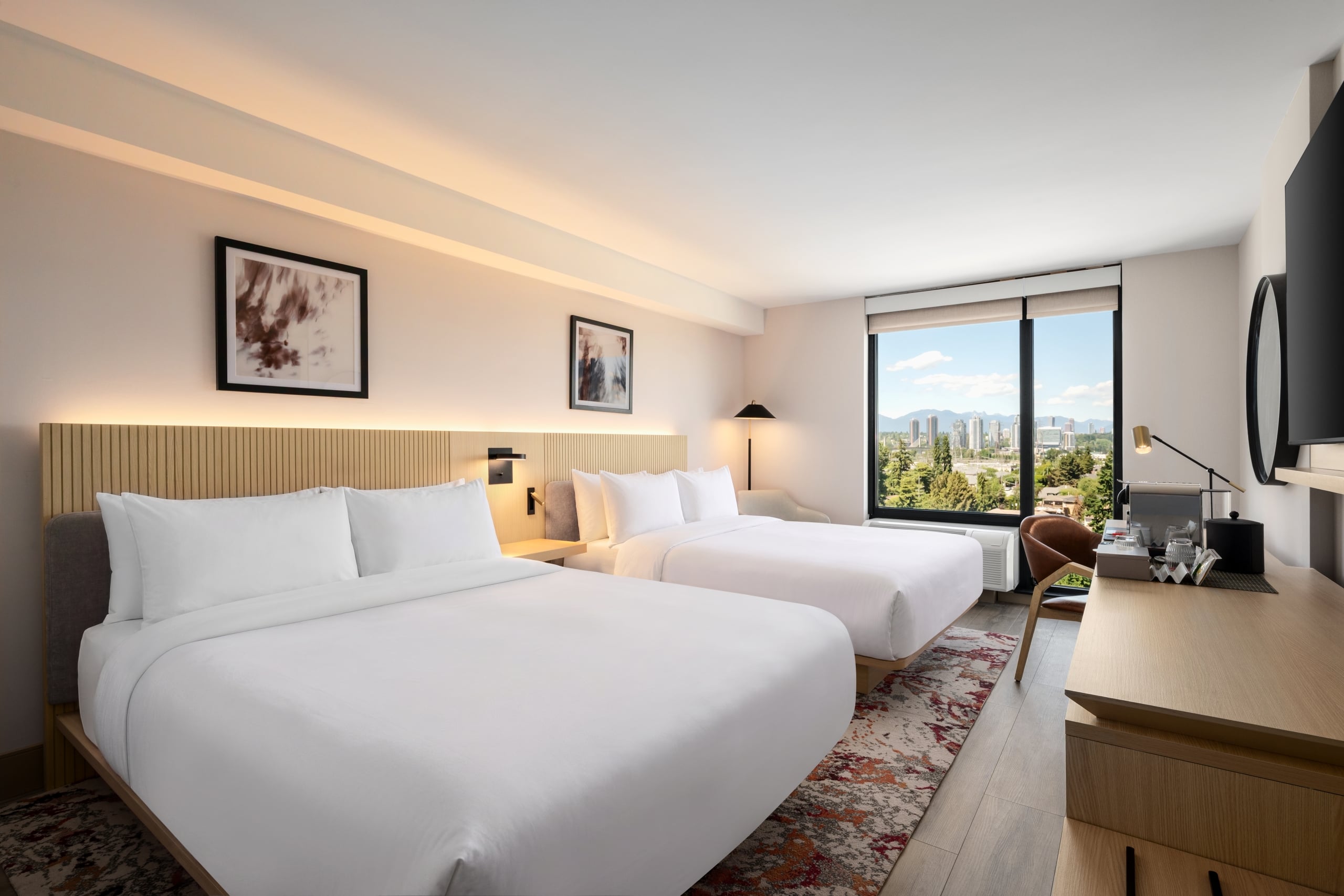 Hotel room with two white-bedding beds, wooden headboard, and a large window overlooking a distant cityscape and hills.