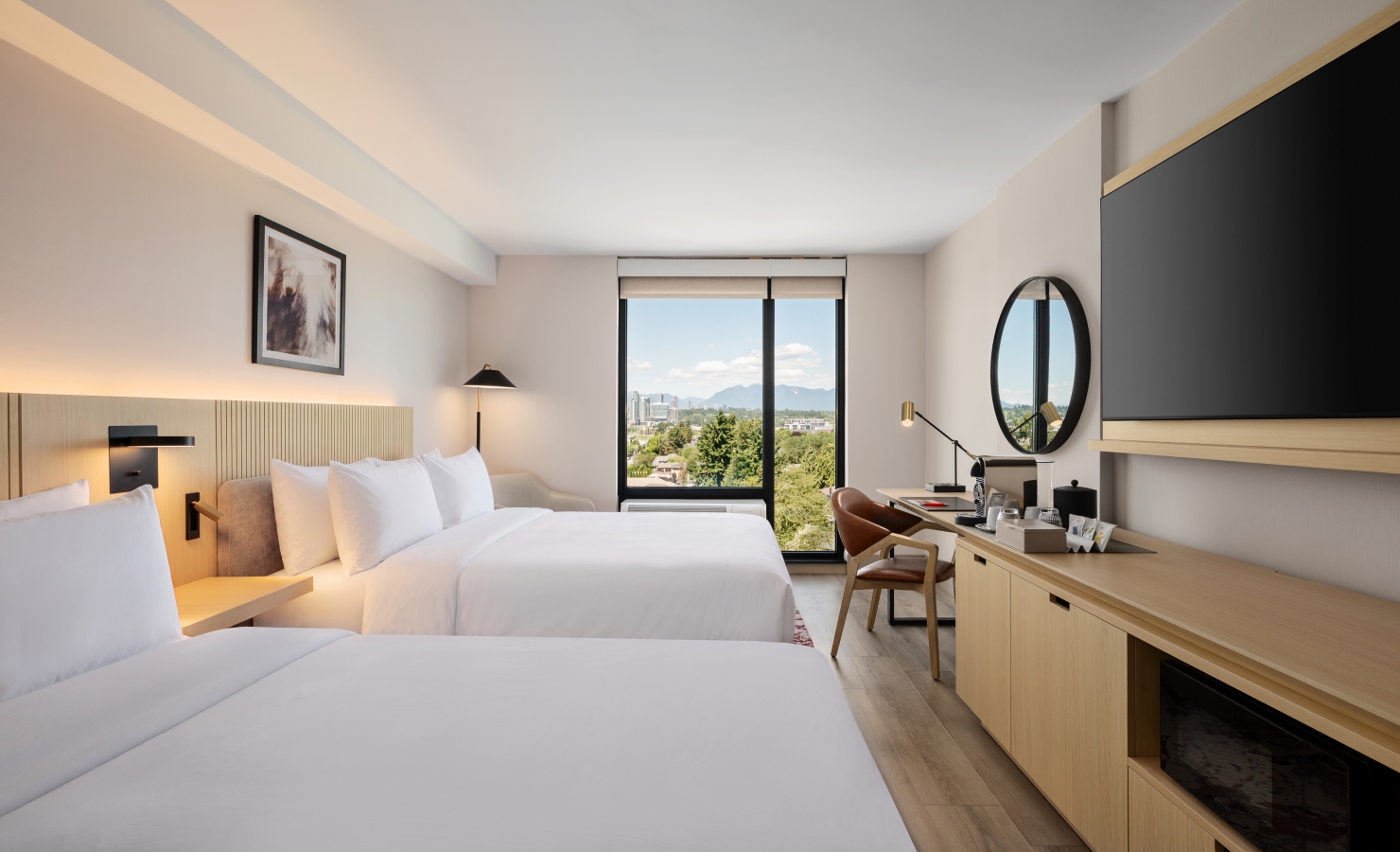 Hotel room with two queen beds, beige headboard, desk, and a round wall mirror overlooking a large window with city and mountain view.
