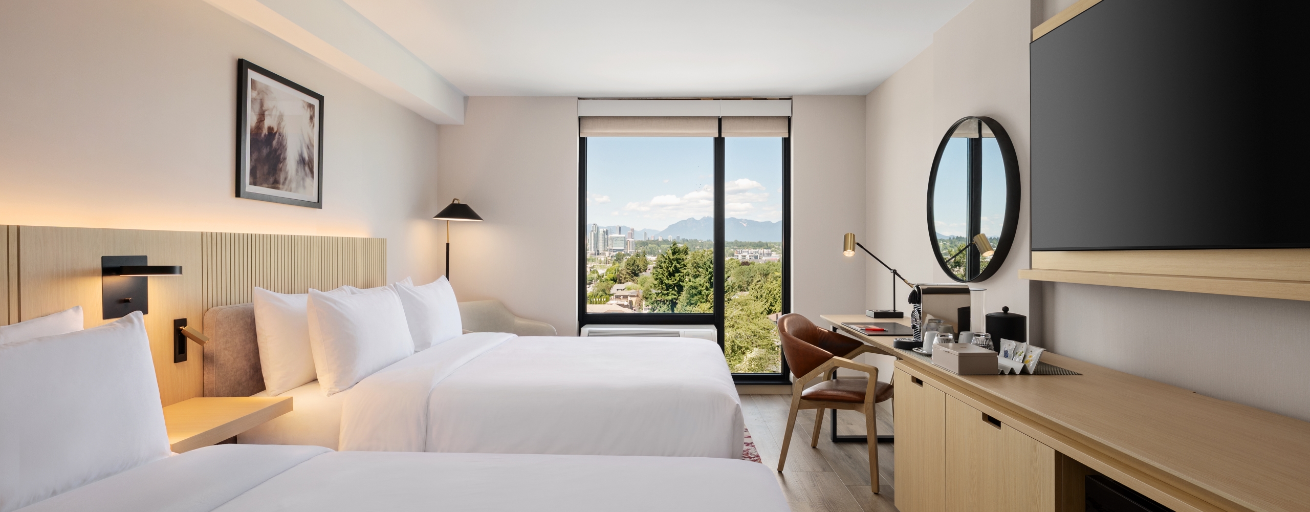 Hotel room with two queen beds, beige headboard, desk, and a round wall mirror overlooking a large window with city and mountain view.